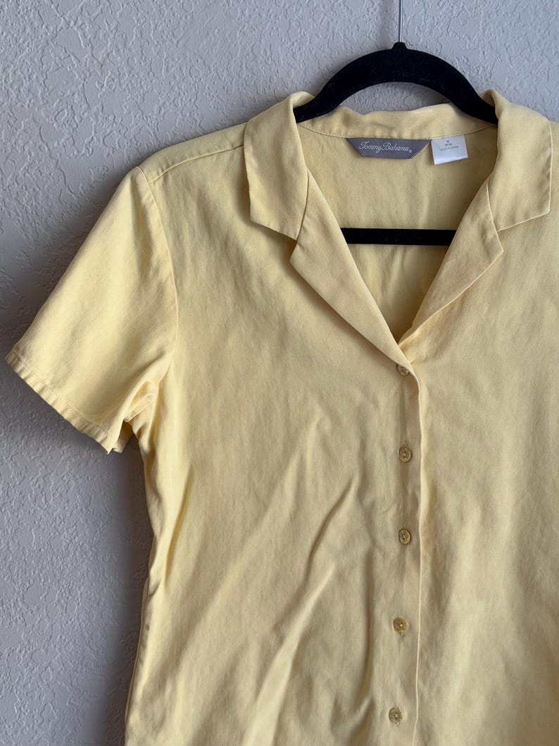 May include: A pale yellow short-sleeved button-down shirt, hanging on a black hanger. The shirt has a collared neckline and a series of small, round buttons down the front. The brand name "Tommy Bahama" is visible on the label.