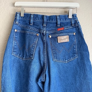 Op de afbeelding: Blauwe denim jeans met het Wrangler-logo op de achterzak. De jeans hangen aan een witte hanger. De achterzakken hebben een kenmerkend W-vormig stiksel. De jeans hebben een klassieke, vintage stijl.