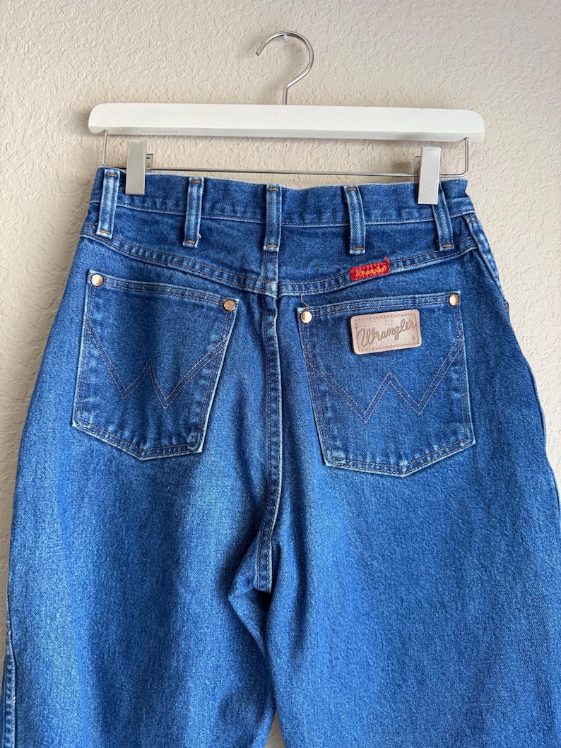 Op de afbeelding: Blauwe denim jeans met het Wrangler-logo op de achterzak. De jeans hangen aan een witte hanger. De achterzakken hebben een kenmerkend W-vormig stiksel. De jeans hebben een klassieke, vintage stijl.