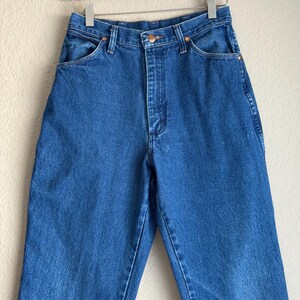 Op de afbeelding: Blauwe denim jeans met een hoge taille, klassiek vijf-zakken ontwerp en een knoop- en ritssluiting. De jeans hangen aan een witte hanger tegen een neutrale achtergrond. De jeans hebben een rechte pijp.