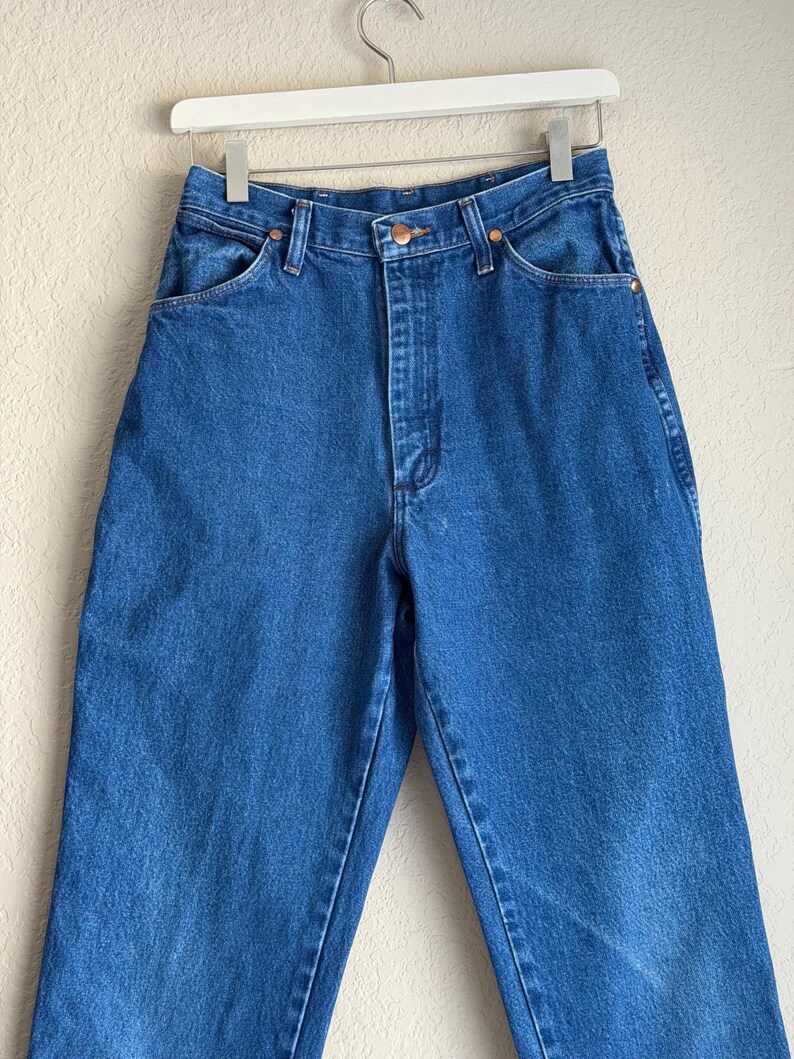 Op de afbeelding: Blauwe denim jeans met een hoge taille, klassiek vijf-zakken ontwerp en een knoop- en ritssluiting. De jeans hangen aan een witte hanger tegen een neutrale achtergrond. De jeans hebben een rechte pijp.