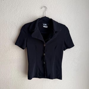 May include: A black short-sleeved button-down shirt with a collar, hanging on a black hanger. The shirt has a fitted silhouette and features small, round buttons down the front. The label reads "I.N."