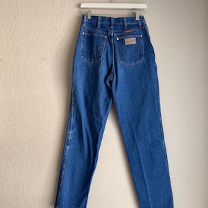 Op de afbeelding: Blauwe Wrangler jeans van denim, hangend aan een witte hanger. De jeans hebben een hoge taille, een klassiek ontwerp met vijf zakken en een Wrangler-patch op de achterzak. De denim heeft een medium wassing met subtiele vervaging.