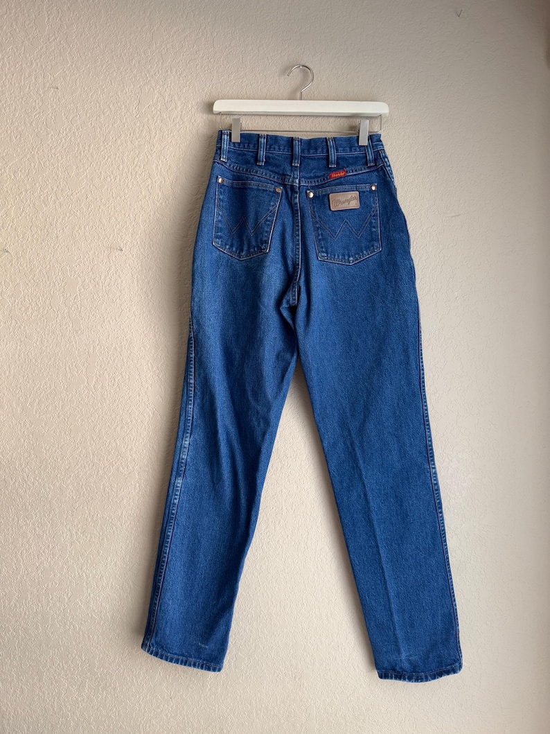 Op de afbeelding: Blauwe Wrangler jeans van denim, hangend aan een witte hanger. De jeans hebben een hoge taille, een klassiek ontwerp met vijf zakken en een Wrangler-patch op de achterzak. De denim heeft een medium wassing met subtiele vervaging.