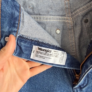 Op de afbeelding: Close-up van een blauwe denim jeans met een rits en een wit label. Het label leest "Wrangler" en bevat maat- en wasvoorschriften. De jeans is gemaakt van 100% katoen.