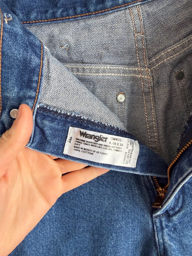 Op de afbeelding: Close-up van een blauwe denim jeans met een rits en een wit label. Het label leest "Wrangler" en bevat maat- en wasvoorschriften. De jeans is gemaakt van 100% katoen.