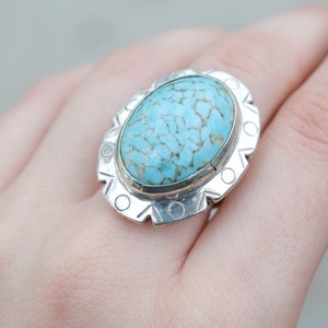 May include: Oval turquoise gemstone ring with a silver setting. The turquoise stone has a light blue color with brown veining. The silver band has a decorative design around the stone.