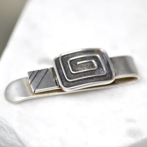 Puede incluir: Pinza de corbata de plata con un dise&ntilde;o cuadrado que presenta un patr&oacute;n de espiral.