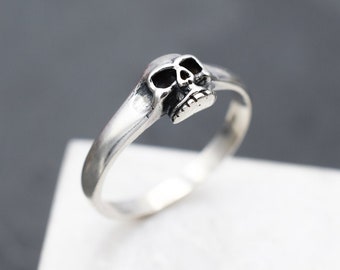 Puede incluir: Un anillo de plata con un pequeño diseño de calavera. La calavera está mirando hacia adelante y tiene un diseño detallado.