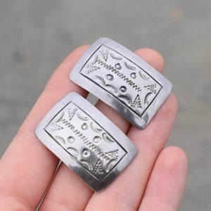 May include: Two rectangular silver-toned metal earrings with rounded corners. Each earring features an engraved design with geometric and organic shapes, including zigzags, dots, and leaf-like patterns. The earrings are held in a person's hand.