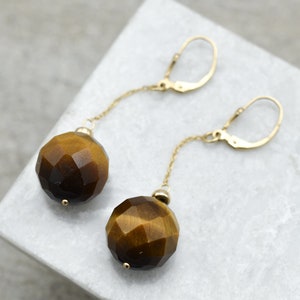 K&ouml;nnte beinhalten: Goldene Ohrringe mit einer langen Kette und einem gro&szlig;en, facettierten Tigerauge-Edelstein.