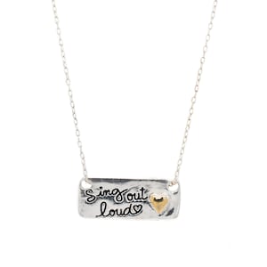 May include: A silver-toned necklace with a rectangular pendant. The pendant has the words "sing out loud" in cursive script, with a small gold-colored heart. The necklace is on a white background.