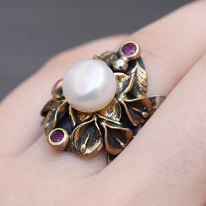 May include: A silver ring with a large white pearl and three small red gemstones set in a floral design.