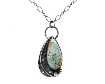 May include: A silver chain necklace with a teardrop-shaped turquoise stone pendant set in a silver frame. The pendant has a textured silver design and small silver beads.