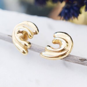 May include: A pair of gold-toned earrings with a wavy, abstract design. The earrings are shown on a wooden surface.