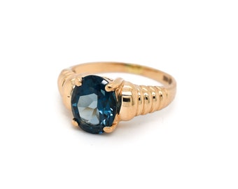 Può includere: Un anello d'oro con una pietra preziosa blu incastonata al centro. La fascia dell'anello è testurizzata con una serie di creste.