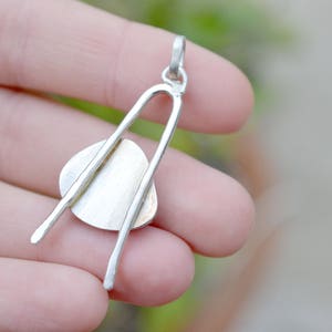 May include: Silver pendant with a round, flat, silver disc attached to two curved silver bars. The pendant is held in a hand.