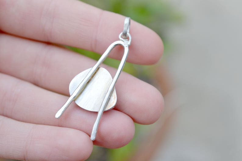 May include: Silver pendant with a round, flat, silver disc attached to two curved silver bars. The pendant is held in a hand.