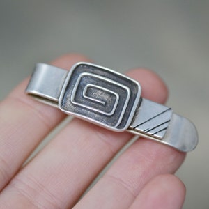 Puede incluir: Pinza de corbata de plata con un dise&ntilde;o geom&eacute;trico. El dise&ntilde;o es un cuadrado con una espiral en su interior.
