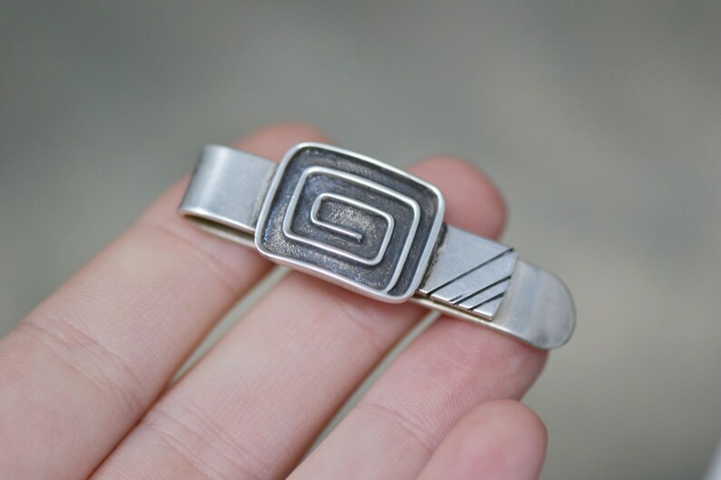 Puede incluir: Pinza de corbata de plata con un dise&ntilde;o geom&eacute;trico. El dise&ntilde;o es un cuadrado con una espiral en su interior.