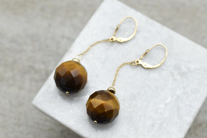 K&ouml;nnte beinhalten: Goldene Ohrringe mit einer langen Kette und einem gro&szlig;en, facettierten Tigerauge-Edelstein.