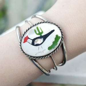 Op de afbeelding: Zilveren armband met een ronde, witte, emaille-achtige bedel met een zwart-witte vogel met een rode kop en een groene cactus. De bedel is omgeven door een zilveren touwachtige rand.