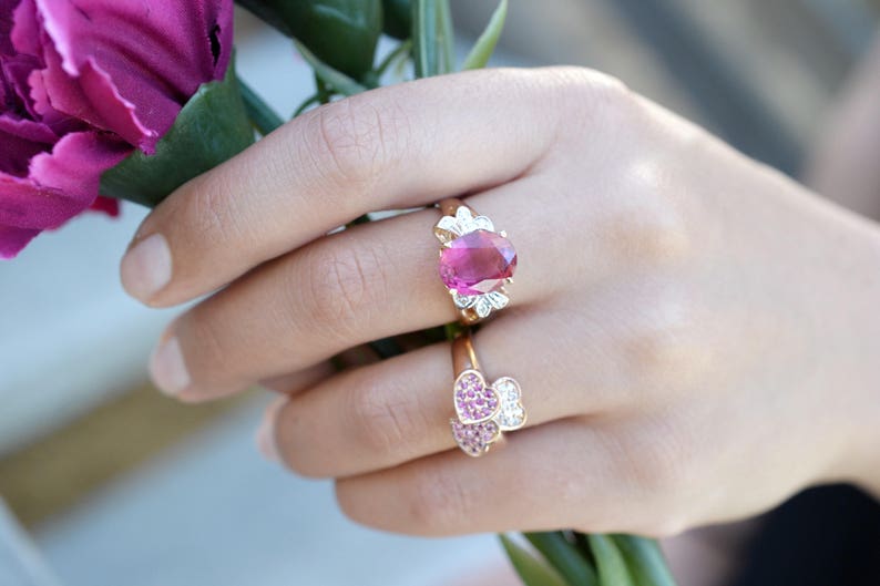 May include: Two rings with pink gemstones. The top ring features a large oval-shaped pink gemstone surrounded by smaller white gemstones. The bottom ring features two heart-shaped pink gemstones.