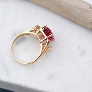 May include: A gold ring with a large, faceted, red gemstone and smaller, clear gemstones. The ring is set on a white marble surface.