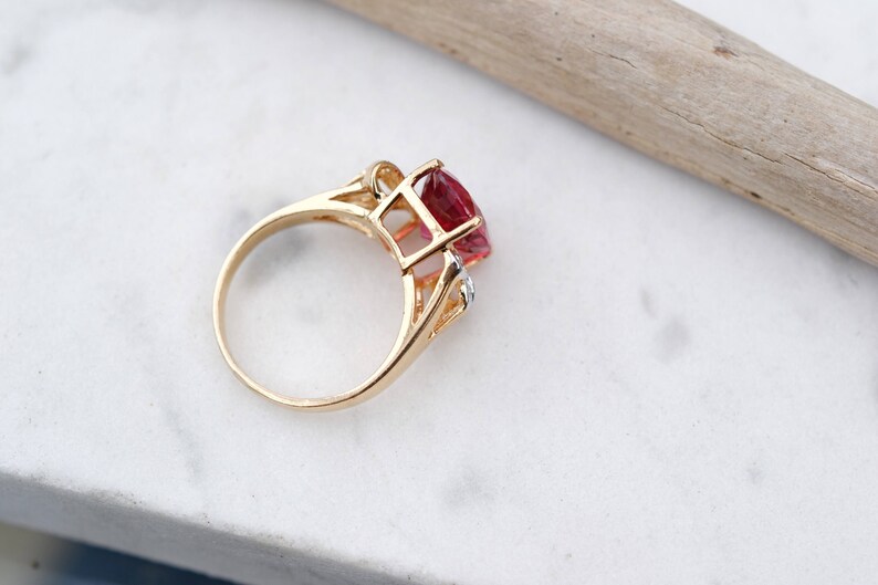 May include: A gold ring with a large, faceted, red gemstone and smaller, clear gemstones. The ring is set on a white marble surface.