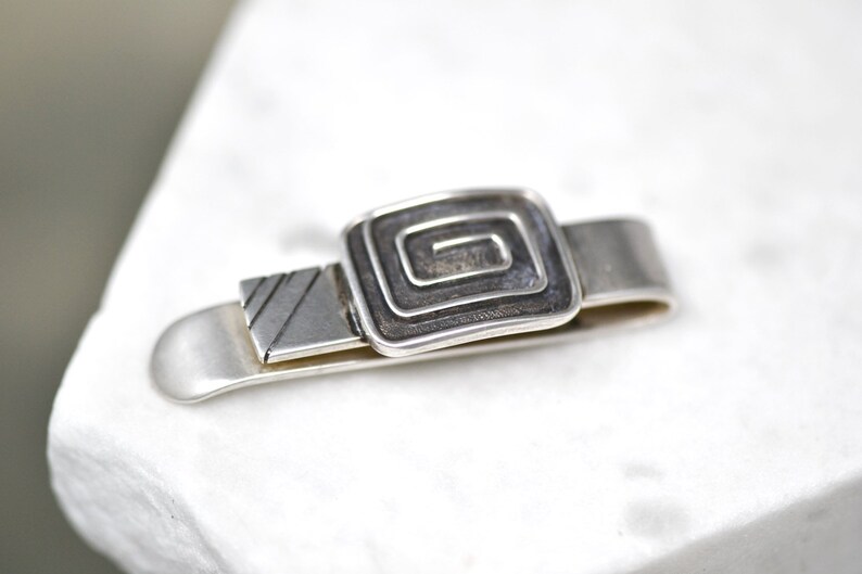 Puede incluir: Pinza de corbata de plata con un dise&ntilde;o cuadrado que presenta un patr&oacute;n de espiral.