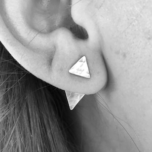 Puede incluir: Un par de pendientes triangulares plateados, uno en la oreja y el otro visible detrás del lóbulo. Los pendientes tienen una textura martillada y la imagen es en blanco y negro.
