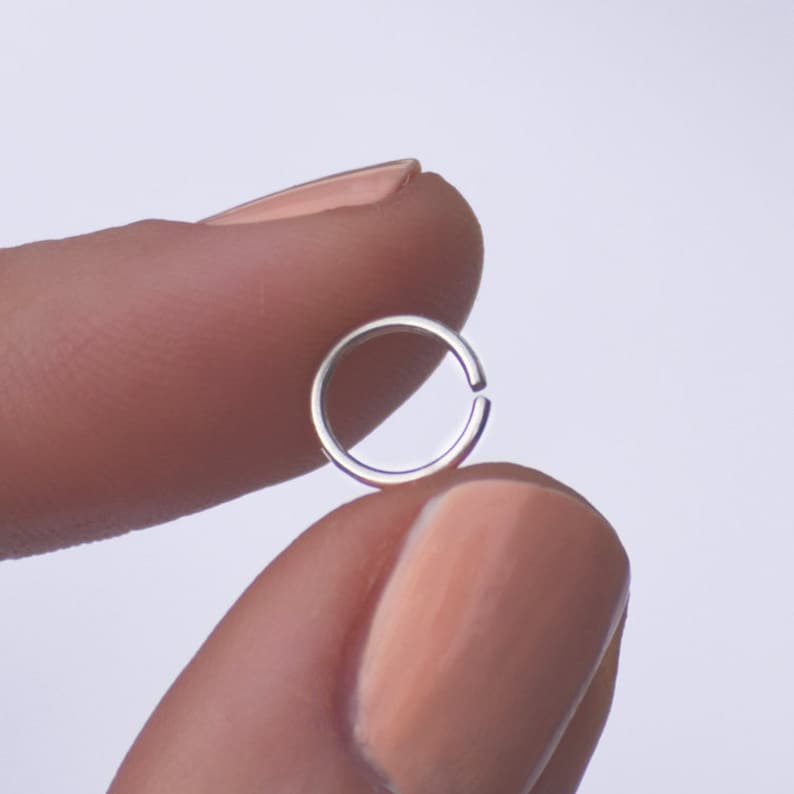 K&ouml;nnte beinhalten: Ein silberner Nasenring, gehalten zwischen zwei Fingern.