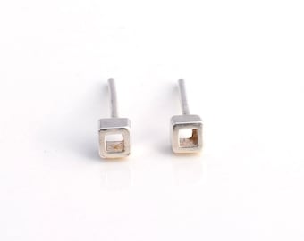 Op de afbeelding: Een paar zilverkleurige oorstekers. Elke oorbel heeft een klein, open vierkant ontwerp aan het einde van een rechte pin. De oorbellen worden gepresenteerd tegen een witte achtergrond, wat hun minimalistische esthetiek benadrukt.