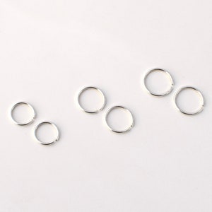 May include: A collection of six small, silver-colored hoop earrings. Each earring is a simple, open circle design, with a small gap for insertion. The earrings are arranged on a white surface, providing a clean and minimalist aesthetic.