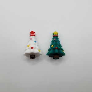 May include: Two miniature Christmas tree ornaments. One is white with a red star and colorful lights. The other is green with a yellow star and colorful lights. Both have brown bases.