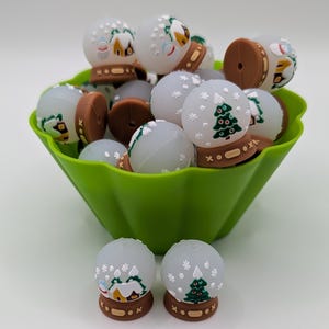 May include: A collection of miniature snow globes with winter scenes, including houses and Christmas trees, displayed in a green bowl. The globes have brown bases and are decorated with white snowflakes. The bowl is a bright green color.