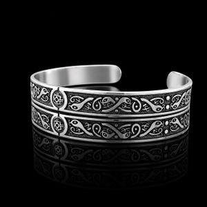 May include: A silver-toned cuff bracelet featuring an intricate, embossed pattern of intertwined serpent-like figures and circular floral motifs. The metal surface has a darkened, oxidised finish within the recessed areas to highlight the design.