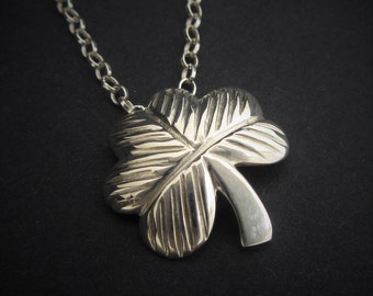 Op de afbeelding: Een zilveren ketting met een vierbladige klaver hanger. De hanger is gedetailleerd met lijnen en heeft een glanzende afwerking.