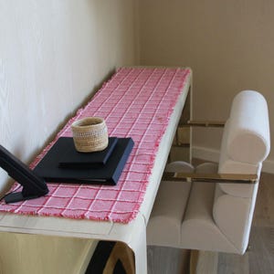 May include: A modern desk setup featuring a red and white checkered table runner, a woven basket, and black notebooks. A white upholstered chair with gold accents sits nearby. The desk is made of light wood.
