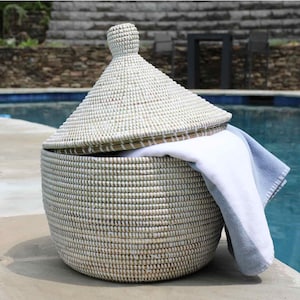 May include: A round, woven basket with a conical lid. The basket is white and beige, with a white towel partially visible inside. The basket is sitting on a concrete surface next to a pool.