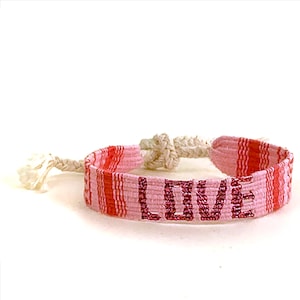 May include: A pink and red woven bracelet with the word "LOVE" spelled out in pink beads. The bracelet has a striped pattern and a knotted closure with a tassel.