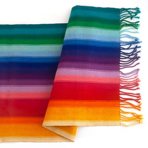 May include: A rainbow striped table runner with fringed edges. The runner is made of woven fabric and features a variety of colors, including red, orange, yellow, green, blue, and purple.