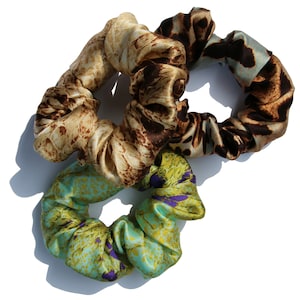 May include: Three patterned scrunchies in brown, beige, and teal with purple accents. The scrunchies are made of silky fabric and are perfect for holding hair in place.