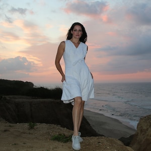 Può includere: Una donna con un vestito bianco si trova in piedi su una scogliera che domina l'oceano al tramonto. Il cielo è un mix di rosa, arancione e blu.