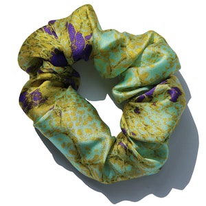 Puede incluir: Un scrunchie con estampado verde y dorado con detalles florales morados. El scrunchie está hecho de una tela suave y sedosa.