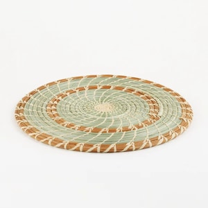 May include: A round woven placemat with a spiral design. The placemat features alternating bands of light green and brown, with a natural fiber border. The center is a lighter beige color. This decorative item is suitable for kitchen and dining decor.