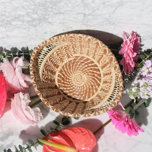 May include: A woven basket with a spiral pattern in shades of brown and beige. The basket is surrounded by pink and white flowers.