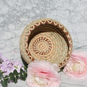 Può includere: Un piccolo cesto rotondo intrecciato con un motivo marrone e verde. Il cesto è fatto di materiali naturali e ha un aspetto delicato e artigianale.