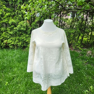 May include: A white lace top with a bell sleeve and a round neckline. The top is on a white mannequin against a green grassy background.
