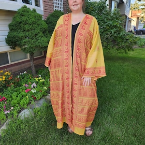 May include: A yellow and red patterned kimono with a black undershirt. The kimono has a long, flowing design and is worn by a person standing in a grassy area.